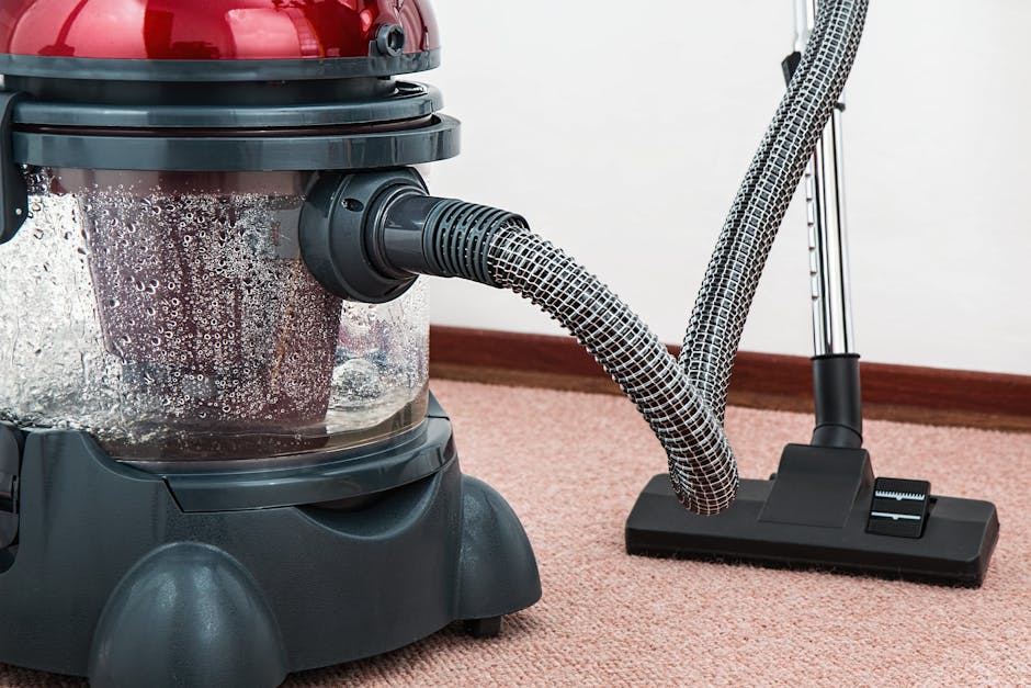 A person wearing full protective gear, including a white suit, face mask, and gloves, is performing deep cleaning on a beige carpet inside a well-lit living room. The individual is using a steam clean