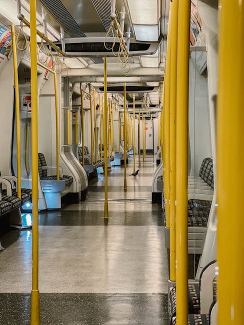Interior of a modern train carriage featuring rows of blue fabric upholstered seats with white plastic backs and blue handrails, arranged in pairs along a central aisle. The carriage is well-lit with